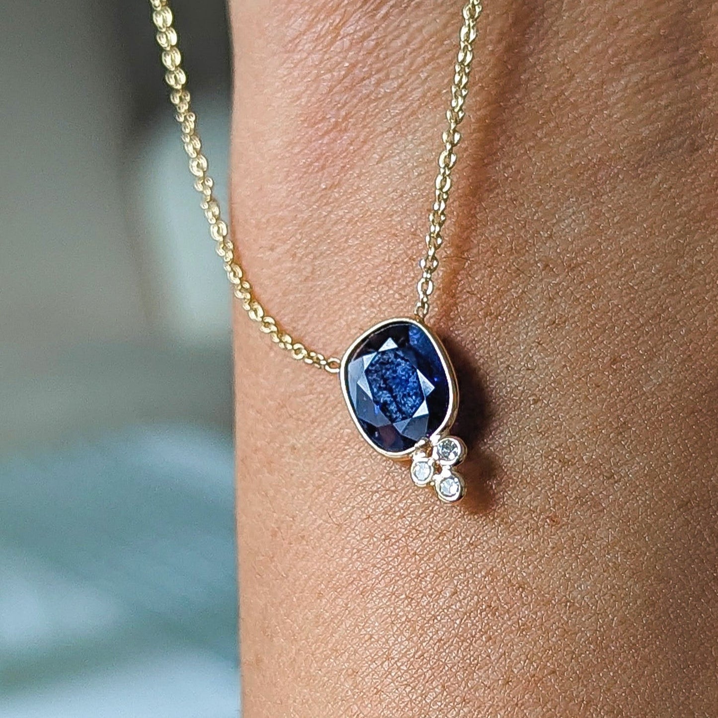 Gold necklace with a blue sapphire gemstone pendant on a person's wrist against a blurred background