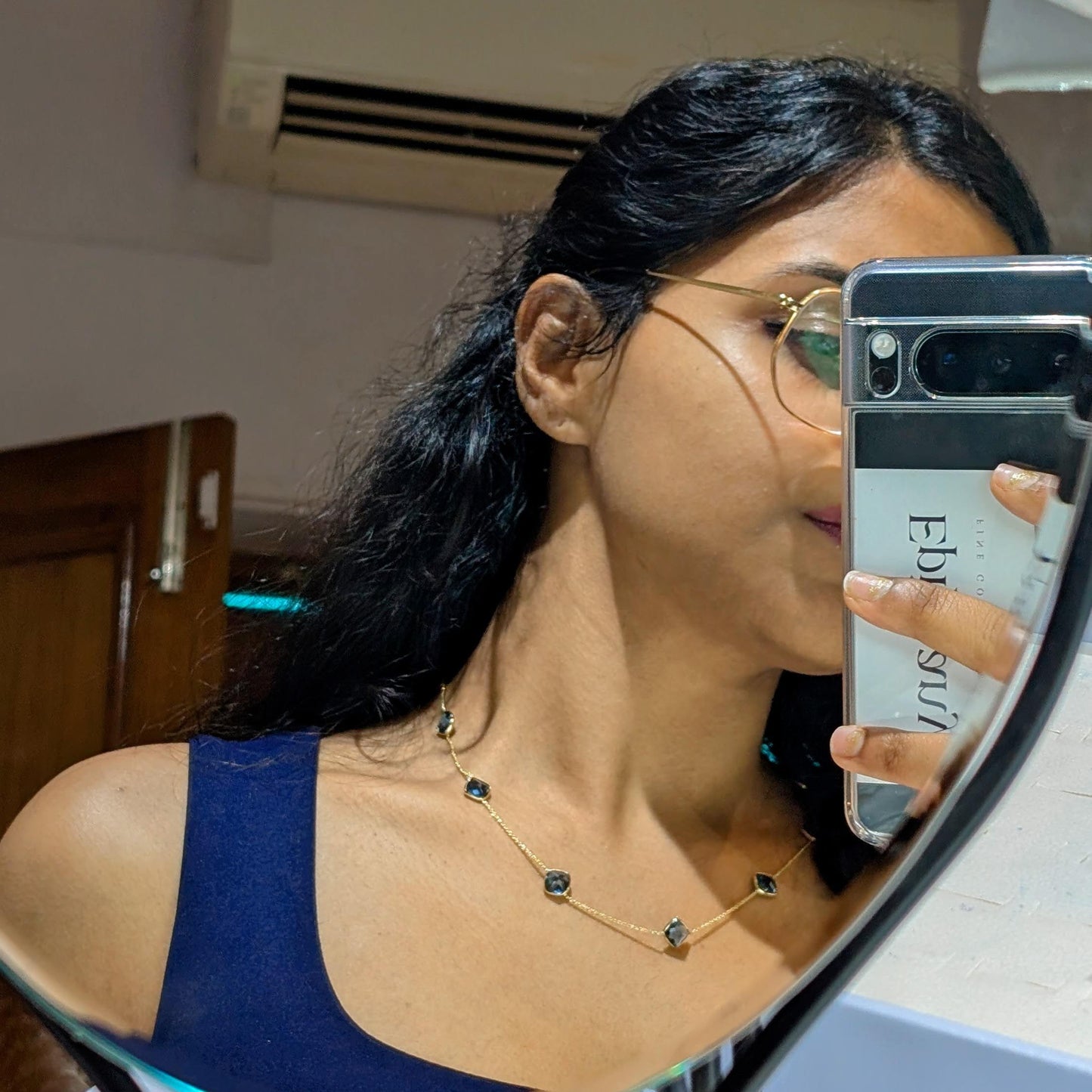 Woman taking a mirror selfie with a phone, wearing glasses and a necklace.