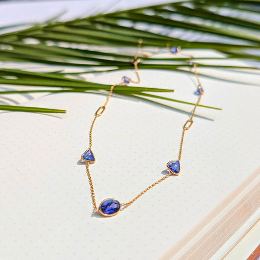 Gold necklace with blue gemstones on a white surface with green leaves in the background