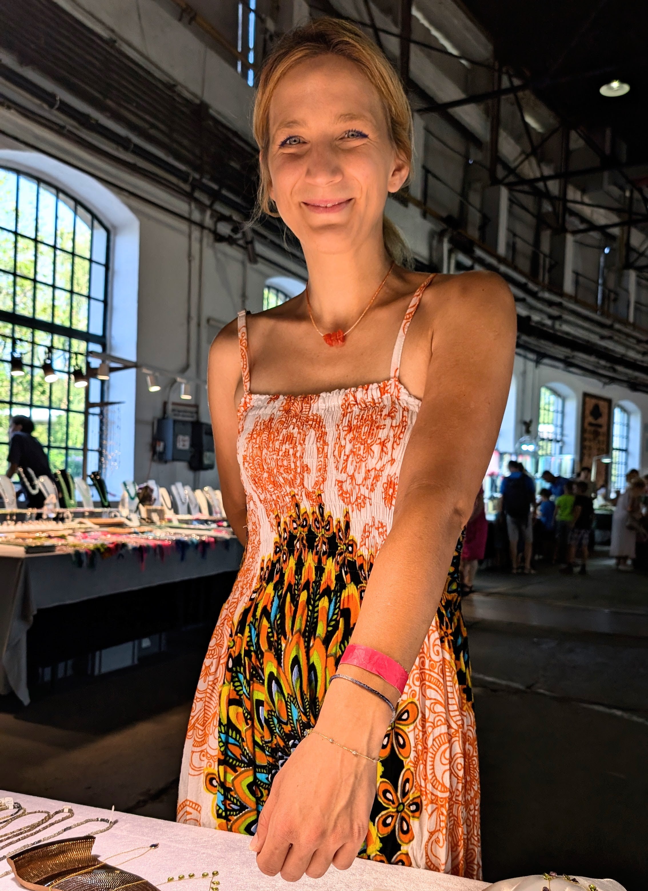 Woman in a colorful dress standing behind a table in a large indoor space with tables and chairs.
