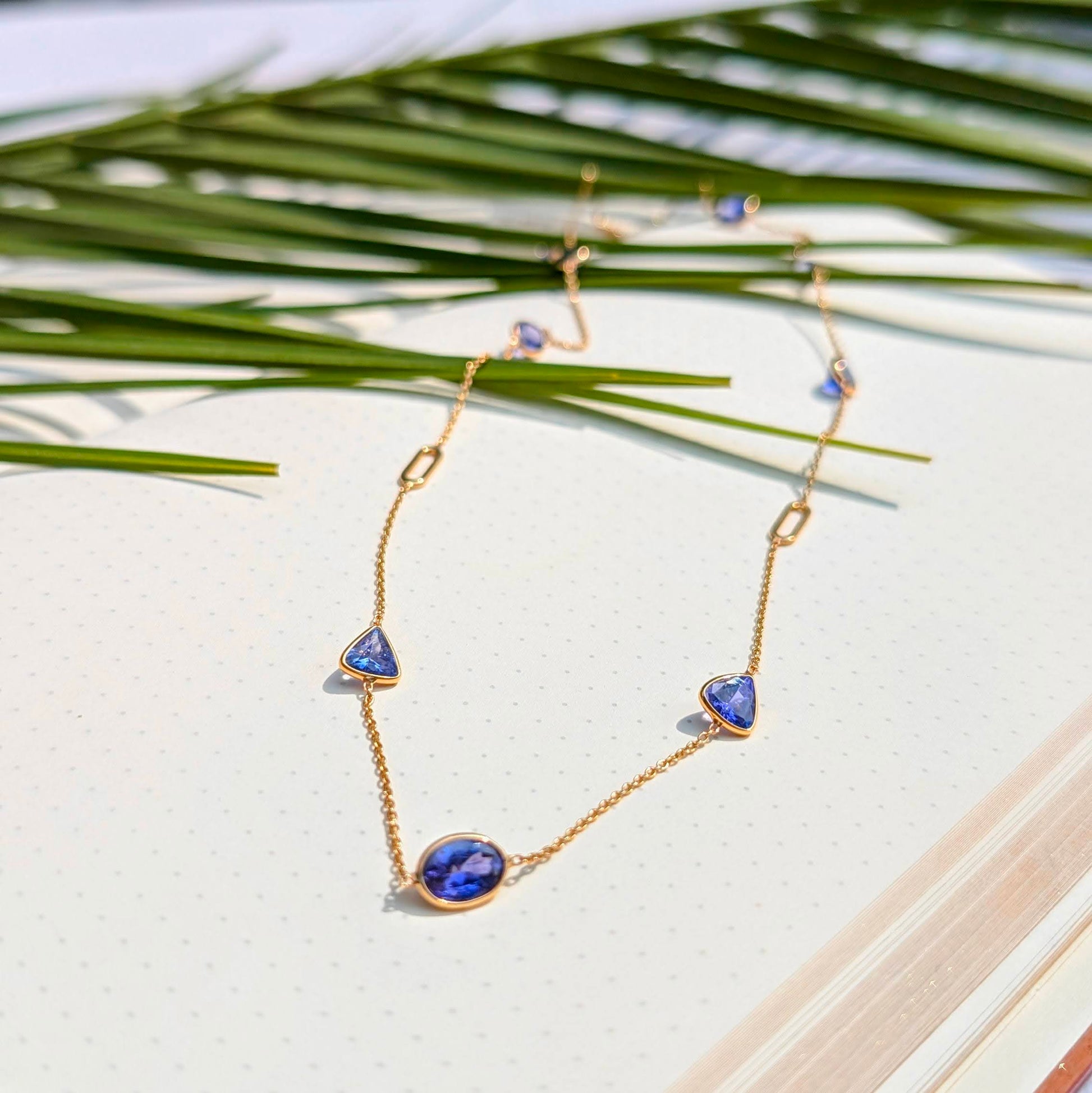 Gold necklace with blue gemstones on a white surface with green leaves in the background