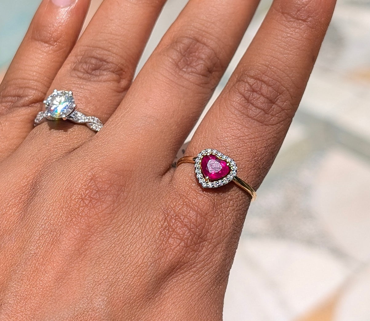 Hand wearing two rings, one with a large diamond and the other with a heart-shaped ruby.